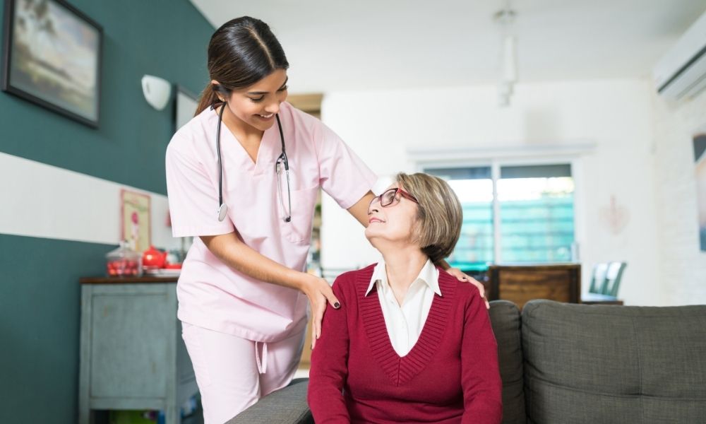 A nurse stands beside a seated older woman and rests a hand on her shoulder.