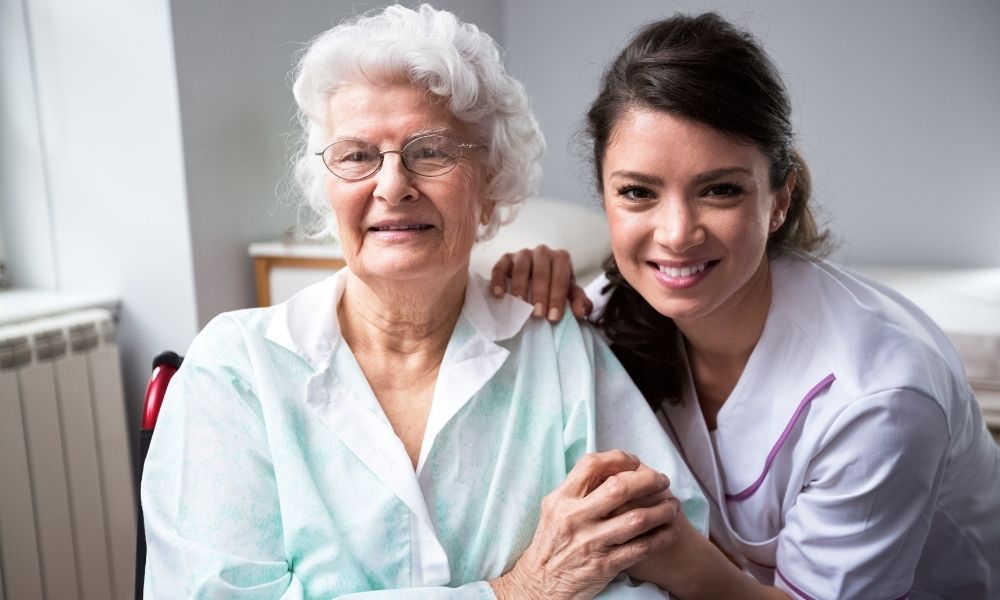 A caregiver sits beside an older woman in a wheelchair and holds her hand.