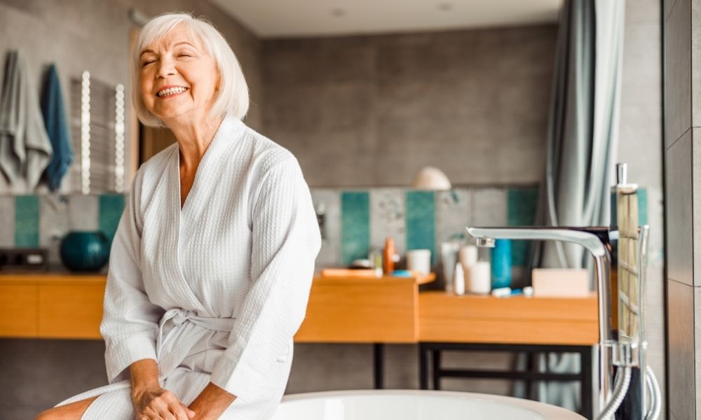 An older woman smiles in a bathrobe in a bright bathroom.
