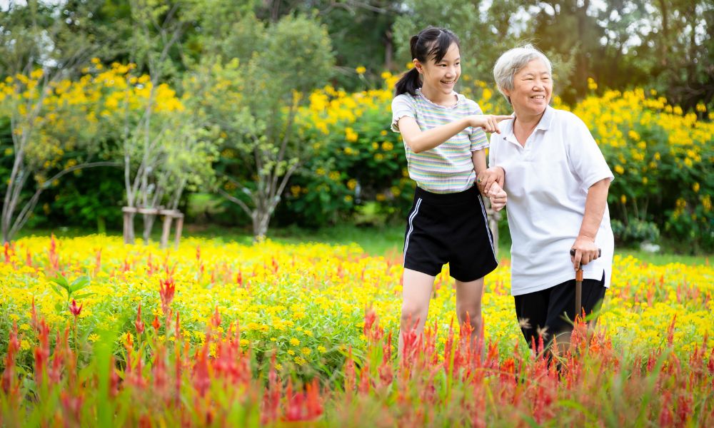 A caregiver walks with an older woman through a flower garden.